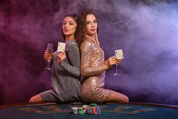 Zwei Mädchen spielen Poker im Casino, halten Champagnergläser und Karten in der Hand und sitzen auf einem Tisch mit Chips darauf. Schwarzer, rauchiger Hintergrund. Nahaufnahme. - Foto, Bild