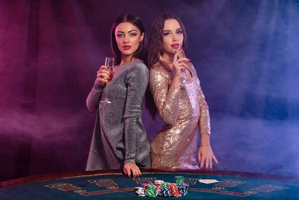 Две девушки играют в покер в казино, держа бокалы шампанского, позируя за столом с фишками, картами на нем. Черный, дымовой фон. Крупный план
. - Фото, изображение