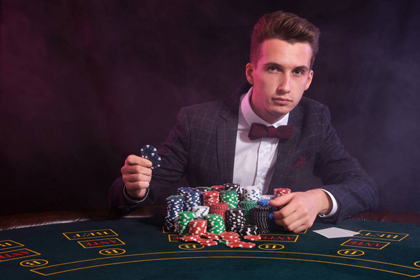 Elegante man speelt poker in het casino. Hij zit aan tafel met stapels gekleurde chips en kaarten erop. zwart, rook achtergrond. - Foto, afbeelding
