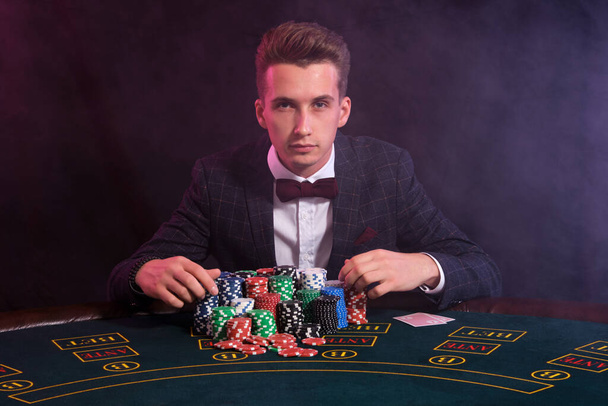 Elegant man is playing poker at casino. He is sitting at the table with stacks of colored chips and cards on it. Black, smoke background. - Foto, immagini
