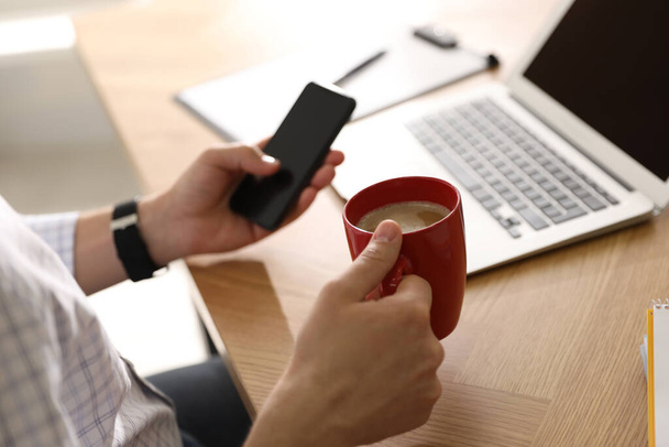 Man with cup of coffee and smartphone at workplace, closeup - Foto, immagini