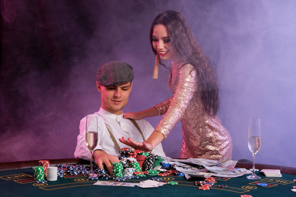 Κομψός άνθρωπος που παίζει πόκερ στο καζίνο συνοδεύεται από ένα κορίτσι. Καθισμένος στο τραπέζι με στοίβες από μάρκες, χρήματα και κάρτες σε αυτό. Μαύρο, φόντο καπνού. - Φωτογραφία, εικόνα