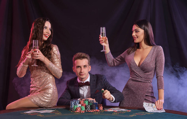 Man die poker speelt in het casino zittend aan tafel met stapels chips, geld, kaarten. Winnen vieren met twee vrouwen. Zwart, rookachtergrond. Close-up. - Foto, afbeelding