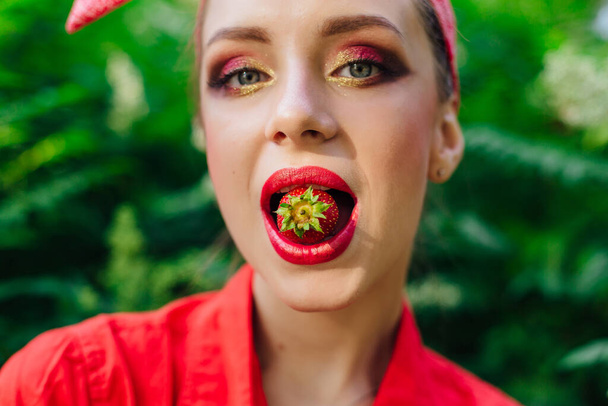Девушка в красной рубашке с ярким макияжем и свежей натуральной клубникой во рту. Закрыть
. - Фото, изображение