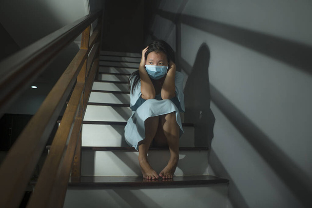 sad portrait of young scared and worried Asian Korean woman in protective mask sitting on stairs at home lockdown and quarantine during covid-19 virus pandemic in dramatic light - Foto, Bild
