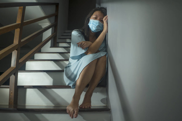 Trauriges Porträt einer jungen verängstigten und besorgten asiatischen Koreanerin in Schutzmaske, die während der Covid-19-Virus-Pandemie in dramatischem Licht zu Hause auf der Treppe sitzt und unter Quarantäne steht - Foto, Bild