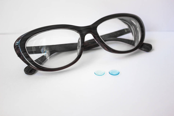 Na bílém pozadí dvě modré čočky pro oči. Za nimi je tmavý ženský rám se silnými brýlemi. Makro fotografie. - Fotografie, Obrázek