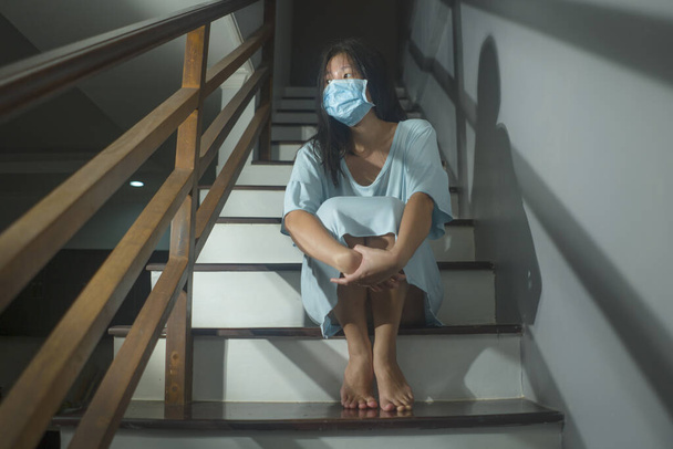 sad portrait of young scared and worried Asian Chinese woman in protective mask sitting on stairs at home lockdown and quarantine during covid-19 virus pandemic in dramatic light - Foto, Bild