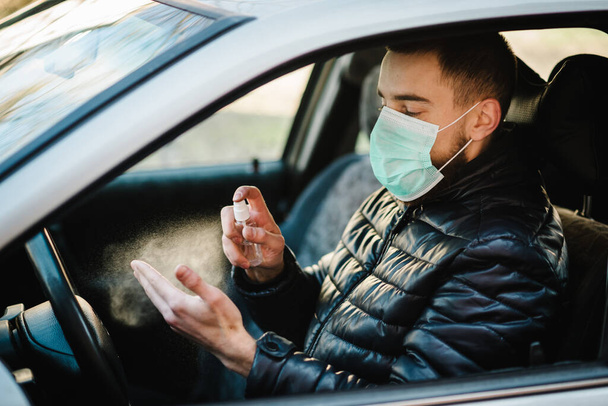 Spraying anti-bacterial sanitizer spray on hand in car, infection control concept. Sanitizer to prevent Coronavirus, Covid-19, flu. Spray bottle. Man wearing in medical protective mask driving a car. - Valokuva, kuva