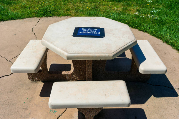 Betonowy stół piknikowy w parku publicznym z powierzchnią niezdezynfekowaną tymczasowy znak ostrzegawczy przymocowany do blatu stołu. - Zdjęcie, obraz
