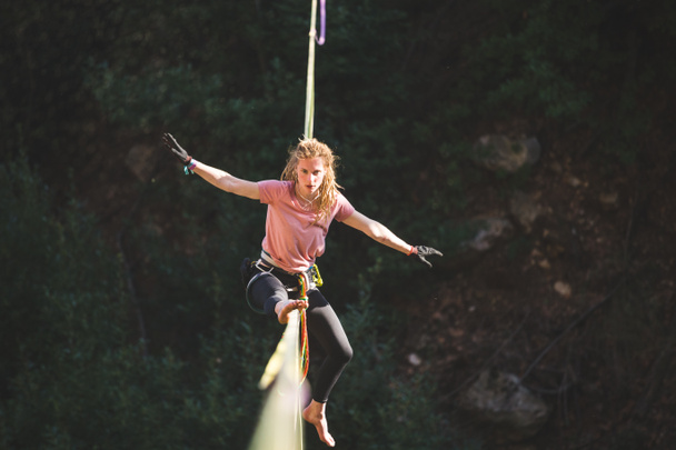 A woman is sitting on a highline, Highliner on a stretched sling, The girl does stretching on a sling stretched over the forest, Trying to get up, highlining, A tightrope walker catches a balance. - Photo, Image