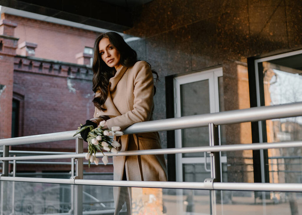 beautiful girl on a walk with flowers laughing smiling, portrait on the street in Sunny weather, with tulips, happy drinking coffee in a coffee shop, reading a book laughing, with makeup in spring summer autumn in gloves in a coat slim model, stylish - Foto, afbeelding