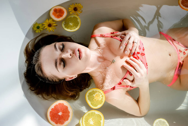 girl in the bathroom sexy water fruit swimsuit underwear bathing hot brunette brown eyes Sunny background sporty seductively wet light chest booty shadows flowers oranges pink splashes photo Studio model fashion glamour hair - Foto, afbeelding