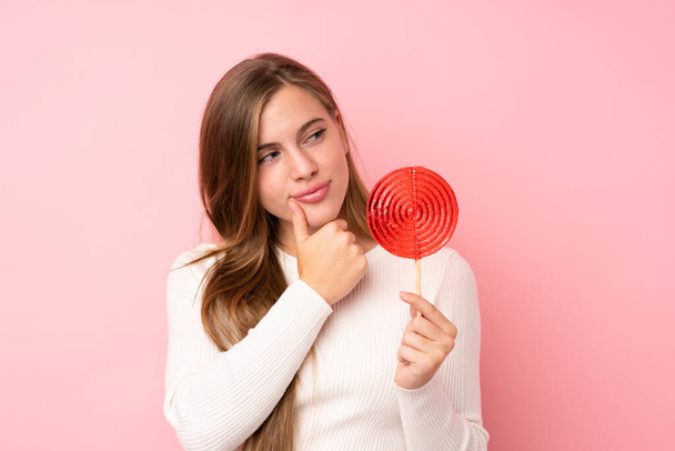Teenager blonde girl holding a lollipop over isolated pink background looking side - Foto, afbeelding