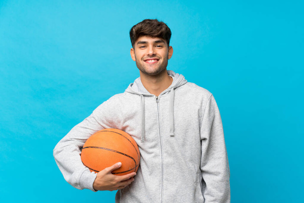 Νεαρός όμορφος άντρας πάνω από απομονωμένο μπλε φόντο με μπάλα μπάσκετ - Φωτογραφία, εικόνα