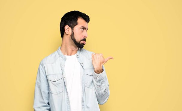 Handsome man with beard unhappy and pointing to the side over isolated yellow background - Photo, Image