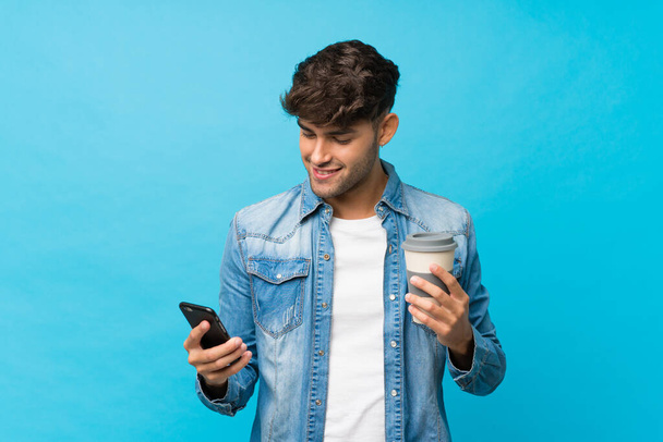 Young handsome man over isolated blue background holding coffee to take away and a mobile - Foto, Imagem