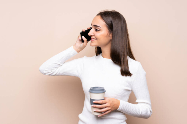 Arka planda izole edilmiş genç bir kadın elinde kahve ve bir cep telefonu tutuyor. - Fotoğraf, Görsel