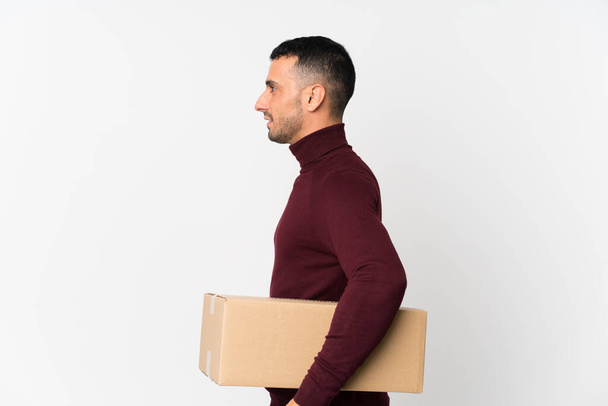 Молодой человек на изолированном белом фоне держит коробку, чтобы переместить ее в другое место в боковом положении
 - Фото, изображение