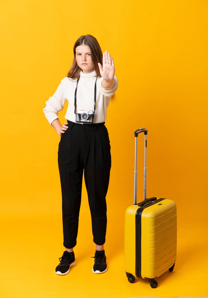 Full body of traveler teenager girl with suitcase over isolated yellow background making stop gesture with her hand - 写真・画像