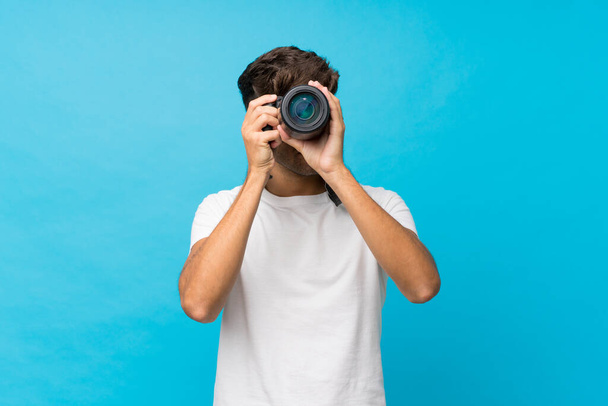 Mladý pohledný muž přes izolované modré pozadí s profesionální kamerou - Fotografie, Obrázek