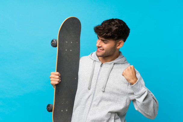 Young handsome man over isolated blue background with skate and making victory gesture - 写真・画像