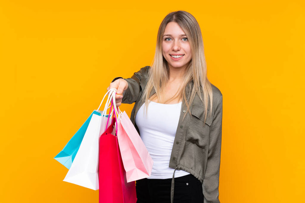 Young blonde woman over isolated yellow background holding shopping bags and giving them to someone - Foto, Bild