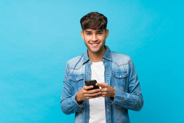 Młody przystojny mężczyzna na odizolowanym tle niebieski wysyłanie wiadomości lub e-mail z telefonu komórkowego - Zdjęcie, obraz