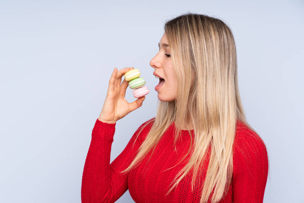 Young blonde woman over isolated blue background holding colorful French macarons and eating it - Photo, Image