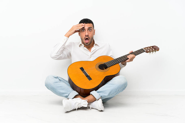 Młody człowiek siedzi na podłodze z gitarą z zaskoczeniem i wstrząśniętym wyrazem twarzy - Zdjęcie, obraz