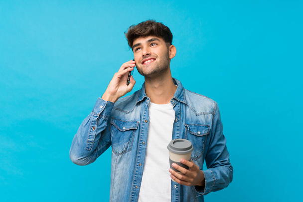 Νεαρός όμορφος άντρας πάνω από απομονωμένο μπλε φόντο κρατώντας καφέ για να πάρει μακριά και ένα κινητό - Φωτογραφία, εικόνα