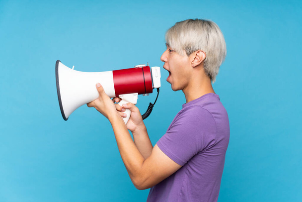 Young asian man over isolated blue background shouting through a megaphone - Foto, afbeelding