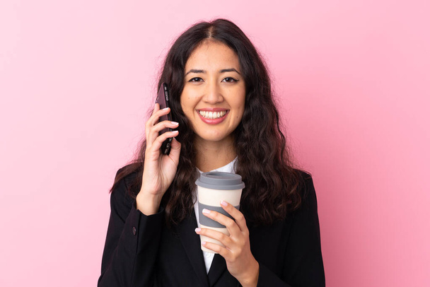 Mieszane wyścigu firma kobieta trzyma kawę do zabrania i rozmowy z telefonu komórkowego na odizolowanym różowym tle - Zdjęcie, obraz
