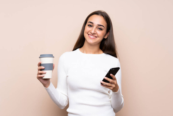 Junge Frau über isolierten Hintergrund mit Kaffee zum Mitnehmen und einem Handy - Foto, Bild