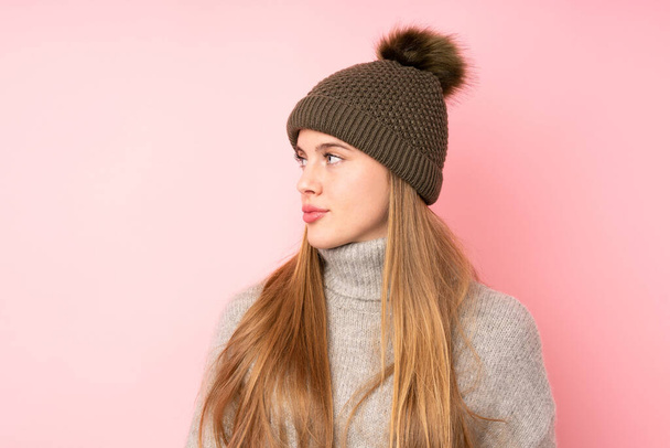 jong tiener meisje met winter hoed over geïsoleerde roze achtergrond kijken kant - Foto, afbeelding