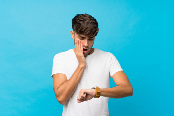 Junger gutaussehender Mann vor isoliertem blauem Hintergrund mit Armbanduhr und überrascht - Foto, Bild
