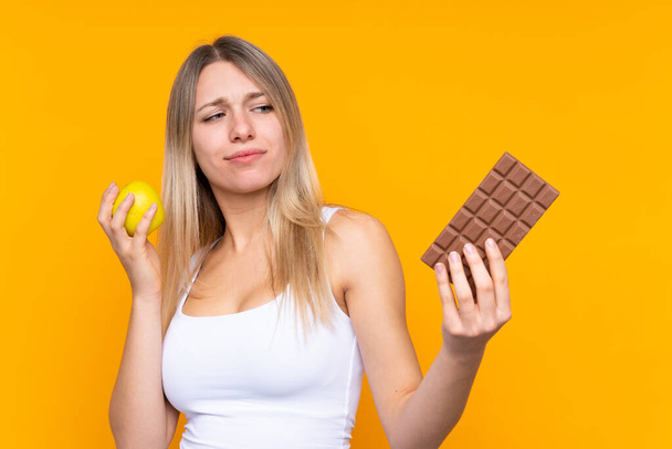 Fiatal szőke nő izolált kék háttérrel, akinek kétségei vannak, miközben az egyik kezében csokoládé tablettát, a másikban almát vesz be. - Fotó, kép