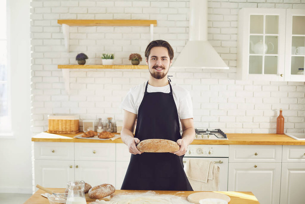 Baker man bearded man holds in his hands fresh bread standing in the kitchen. - Fotografie, Obrázek