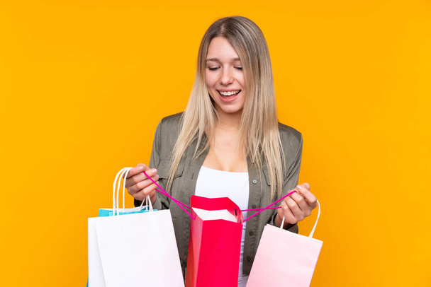 Mujer rubia joven sobre fondo amarillo aislado sosteniendo bolsas de compras y mirando dentro de ella
 - Foto, imagen