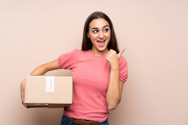 Young woman over isolated background holding a box to move it to another site and pointing side - Photo, Image