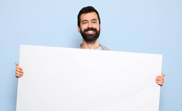 Happy Handsome man with beard holding an empty placard over isolated blue background - Fotoğraf, Görsel