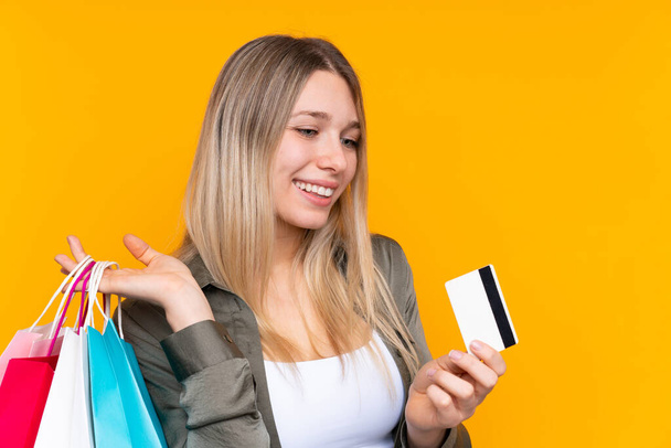 Young blonde woman over isolated yellow background holding shopping bags and a credit card - Foto, Imagem