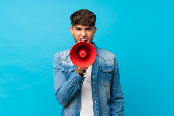 Молодий красивий чоловік над ізольованим синім тлом кричить через мегафон
 - Фото, зображення
