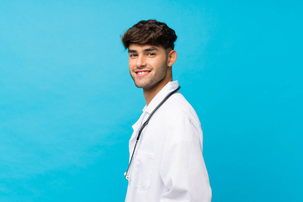 Молодой красивый мужчина на изолированном синем фоне в халате врача
 - Фото, изображение
