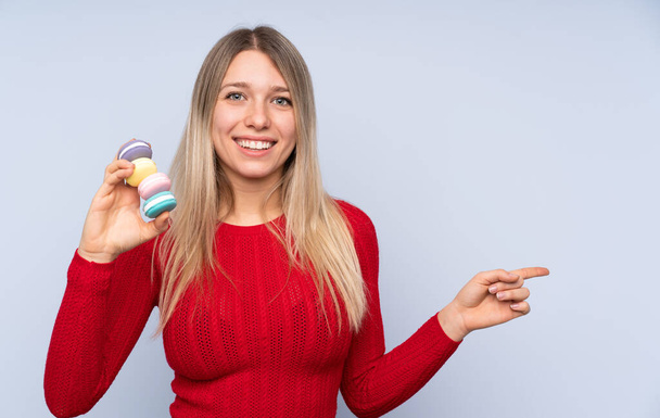 Young blonde woman over isolated blue background holding colorful French macarons and pointing side - Photo, Image
