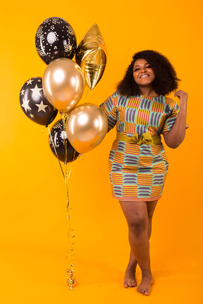 Vakantie, verjaardagsfeest en leuk concept - Portret van lachende jonge Afro-Amerikaanse jonge vrouw die er lief uitziet op gele achtergrond met ballonnen. - Foto, afbeelding