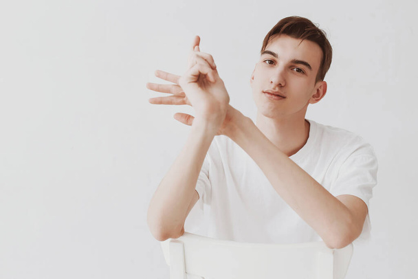 Portrait of a beautiful young guy in a white T-shirt sitting against a white wall and running on a chair, rubbing his hands. - Фото, зображення