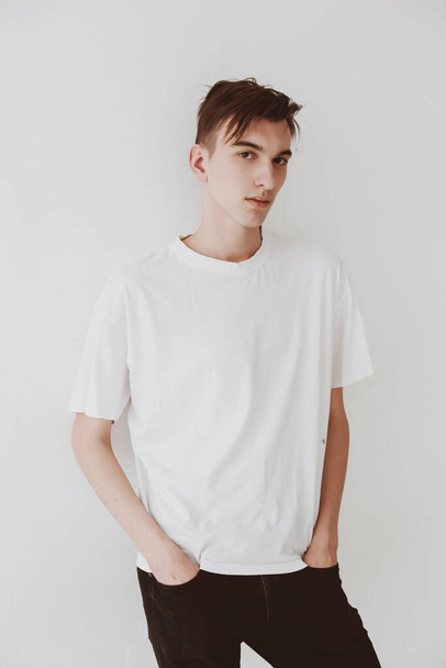 A young, handsome guy, in a white T-shirt, leaned against the white wall. Hands in the pants of the pants. Student, life style. - Foto, afbeelding