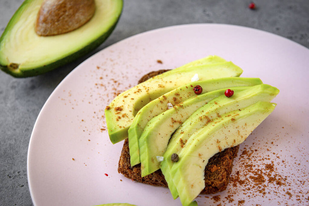 sandwich ,  few slices of avocado with spices on a piece of bread on a plate on a gray background, half a fresh avocado - Φωτογραφία, εικόνα