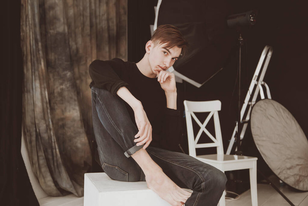 A handsome male model, in a photo studio. On the background of studio equipment. A guy barefoot, in jeans and a black sweater. Sits on a white cube. - Foto, Bild